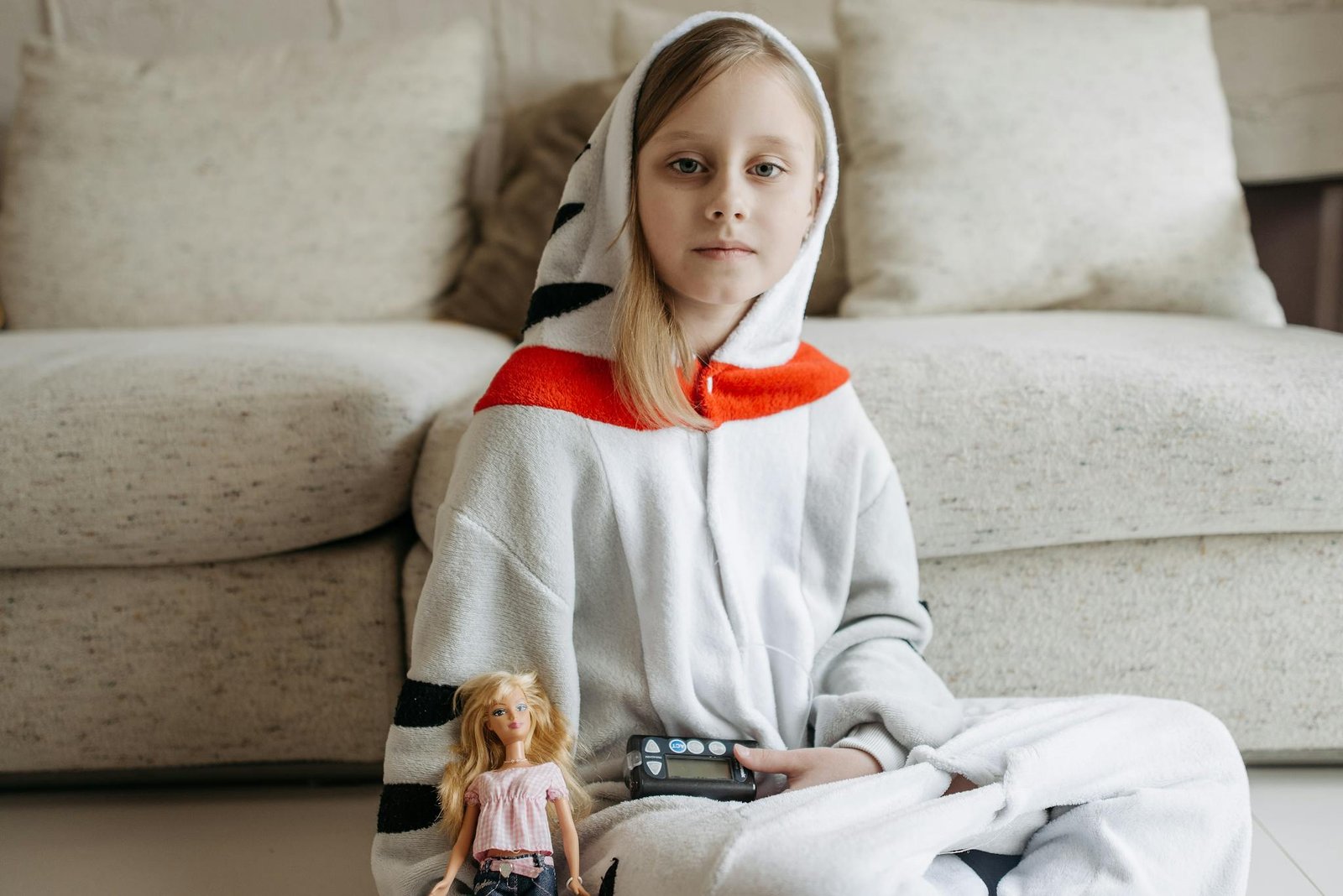 Young girl in onesie sitting at home with an insulin pump and doll, illustrating diabetes management.